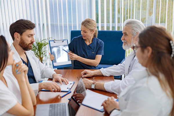 A multidisciplinary tumor board sits at a rectangular table reviewing a mesothelioma case. The board includes a thoracic surgeon, a radiologist, a pathologist and a medical oncologist.