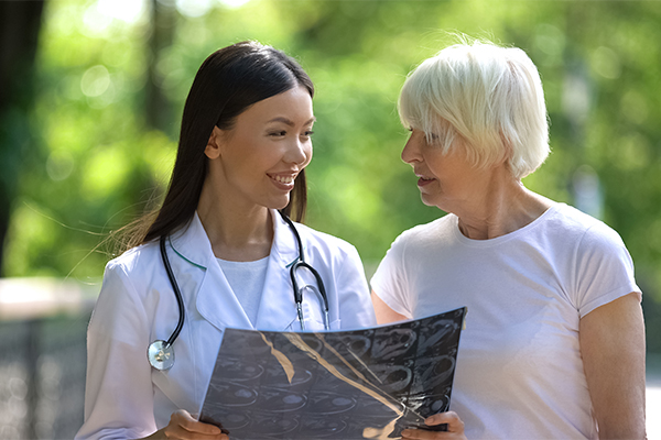 An smiling doctor is holding an image scan. Her patient is an older woman with short white hair, standing to the right.